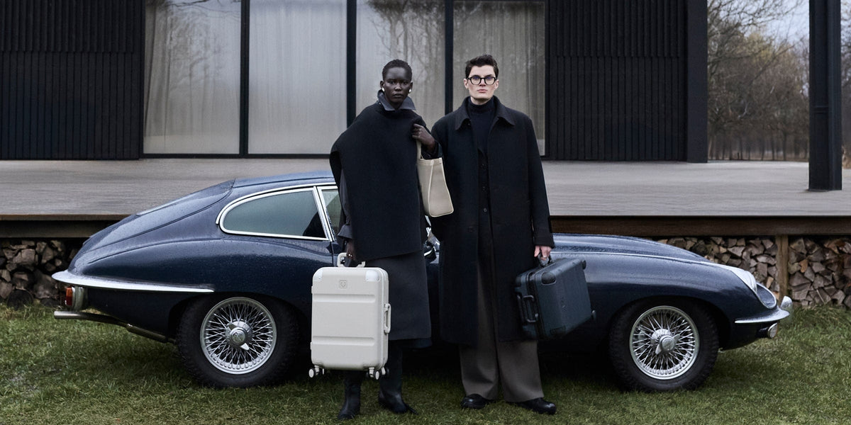 a couple of people in black coats holding luggage and standing in front of a car