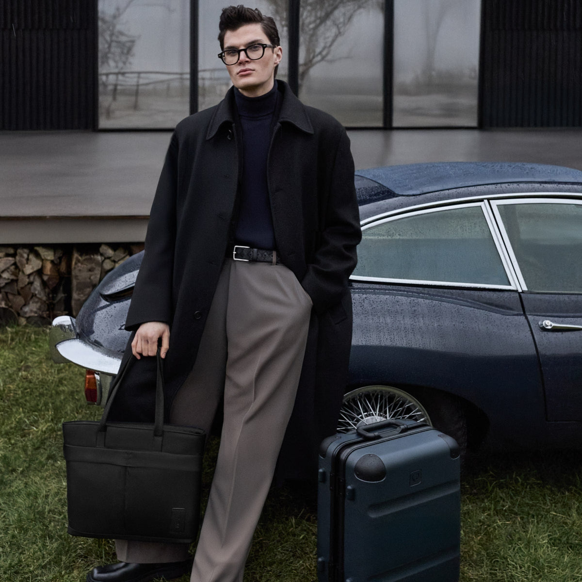 a man in a coat and glasses leaning against a car